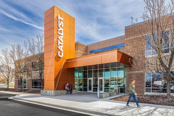 Modern building with a prominent orange sign reading "CATALYST." Glass entrance, people sitting and walking, bare trees, sunny day, and a dynamic atmosphere.