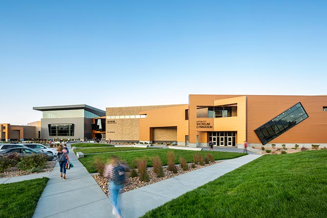 Modern school building with a warm exterior, large windows, and geometric design. Students walk on paths amidst green lawns under a clear blue sky.