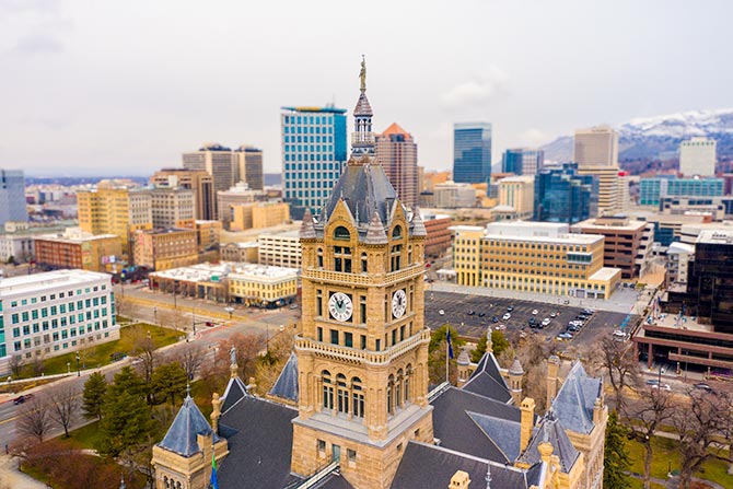AIA Utah Fellows 1889-2026; Aerial view of a historic clock tower with intricate architecture in the foreground, surrounded by an urban skyline of modern buildings and snow-capped mountains in the background. The scene conveys a blend of historical and contemporary urban life.