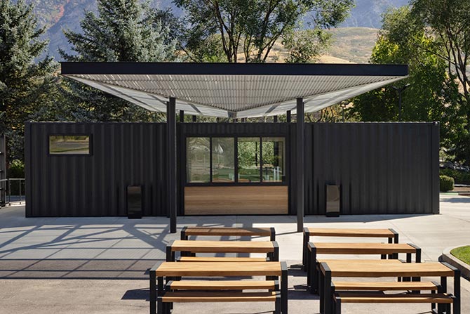 2025 Small Project Honor Award: Scratch Clubhouse; Modern outdoor pavilion with a black metal and wood structure. Wooden tables and benches sit on a concrete patio, surrounded by trees and mountains.