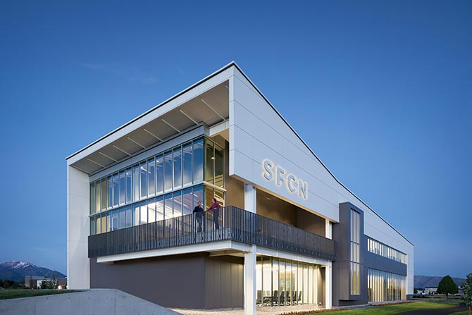 2025 Architecture Honor Award: Spanish Fork Community Network; Modern building with "SFGN" sign, large glass windows, and an upper balcony under a clear blue sky. Two people stand on the balcony, creating a professional tone.