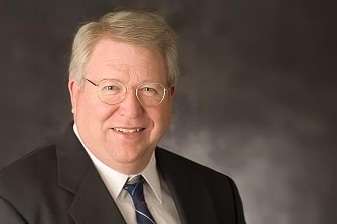 In Memoriam: Stephen Martin Ehninger; Elderly man with glasses smiles warmly, wearing a suit and tie, against a dark, blurred background, conveying a professional and welcoming tone.