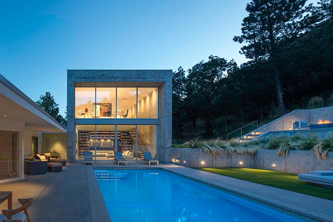 2025 Small Project Merit Award: The Cube; Modern house with large glass windows beside a lit swimming pool at dusk. Surrounded by trees, the scene is serene and features patio seating.