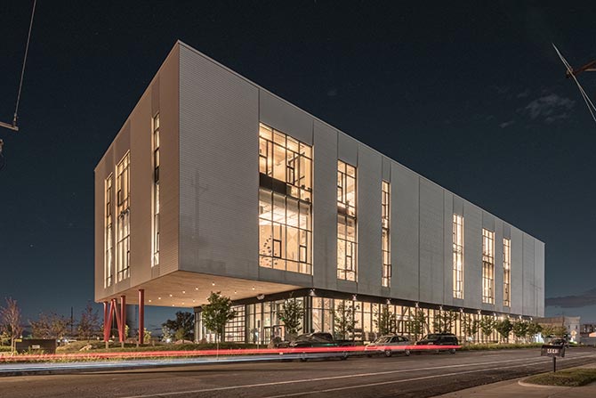 The Front at South Main; Modern building with large, illuminated windows at night. It's elevated on red pillars with cars passing in front, conveying a dynamic, urban atmosphere.