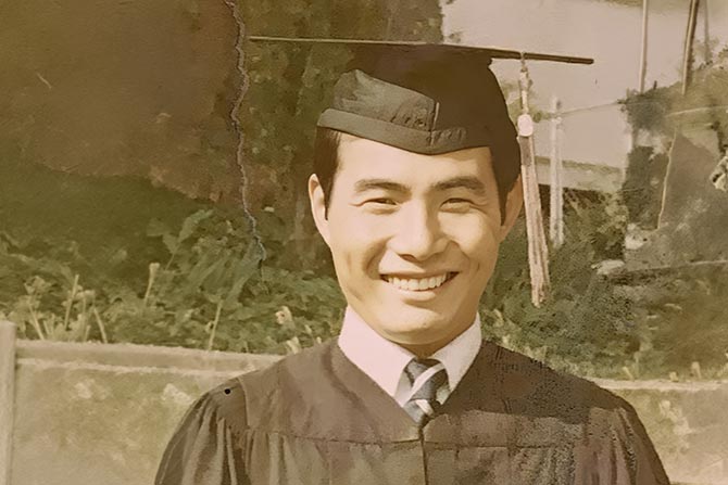 In Memoriam: Tony Tsunehisa Yamada; A man smiling in a graduation gown and cap stands outside. Sunlight highlights his face against a blurred background of greenery and stone.