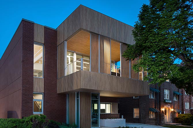 Utah Clean Energy Climate Innovation Center; Modern building at dusk, featuring a blend of brick and wood textures, large windows glowing with interior light and a tree framing the right side.